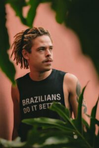 Confident man with dreadlocks and tattoo in sleeveless shirt stands outdoors.