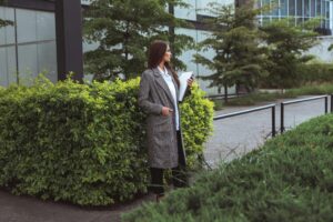 A woman in a coat stands outdoors with documents, surrounded by greenery.