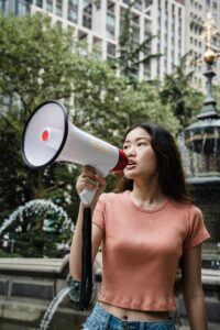 Woman holding a megaphone at an urban protest, surrounded by city buildings and fountain.